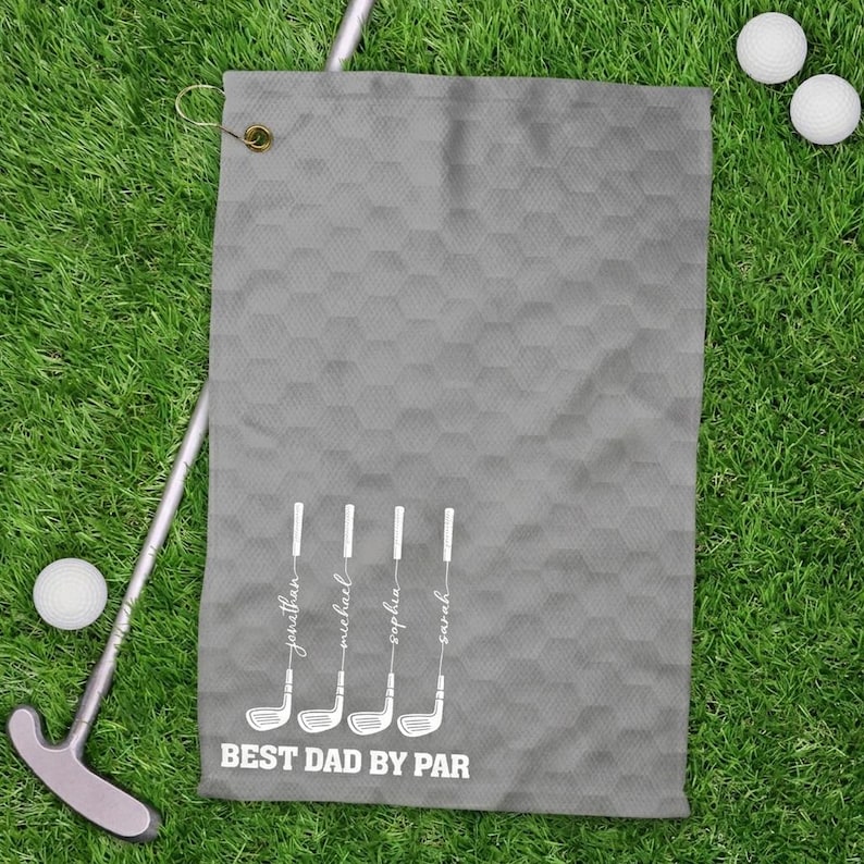 May include: A grey golf towel with a honeycomb pattern, golf clubs, and the text "BEST DAD BY PAR." The names Jonathan, Michael, Sophia, and Sarah are printed on the golf clubs. Two golf balls and a putter are on the green grass.