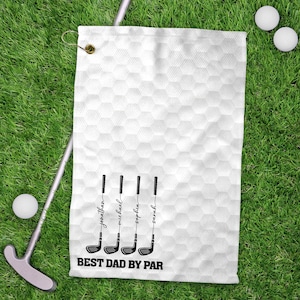 May include: White golf towel with a honeycomb pattern, featuring four golf club illustrations with names and the text "BEST DAD BY PAR". The towel is on green grass, with golf balls and a putter visible.
