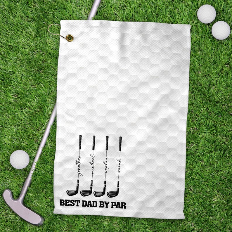 May include: White golf towel with a honeycomb pattern, featuring four golf club illustrations with names and the text "BEST DAD BY PAR". The towel is on green grass, with golf balls and a putter visible.