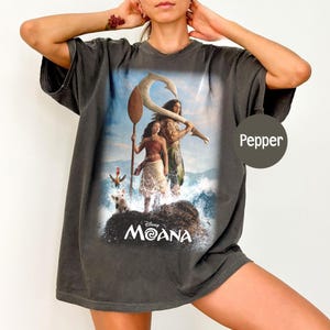 K&ouml;nnte beinhalten: Dunkelgraues T-Shirt mit einem lebendigen Grafikdesign aus dem Disney-Film Moana. Das Design zeigt Moana, Maui und Pua das Schwein an einem felsigen Ufer mit Meereswellen. Das Shirt hat eine lockere Passform und einen V-Ausschnitt.