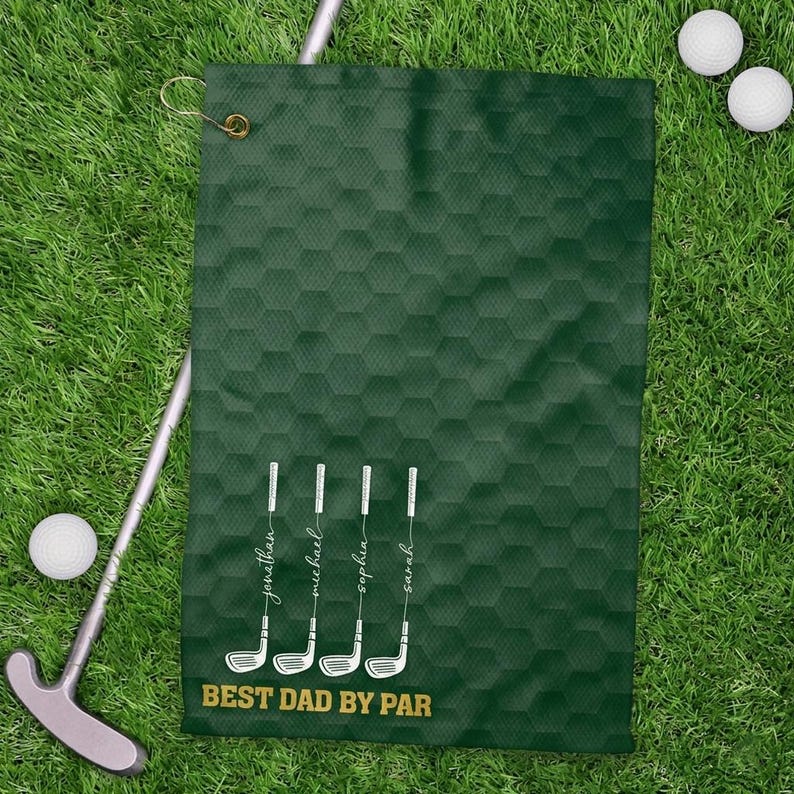 May include: A dark green golf towel with a honeycomb pattern and the text "BEST DAD BY PAR". The towel features white golf club illustrations with names below them. Golf balls and a putter are visible on the grass.