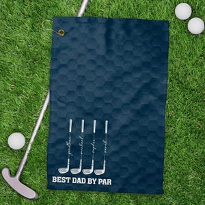 May include: Navy blue golf towel with a honeycomb pattern. Features white golf club illustrations with names and the text "BEST DAD BY PAR". Includes a brass grommet for hanging. Golf balls and a putter are also visible.