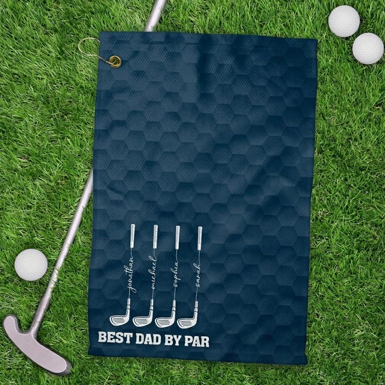 May include: Navy blue golf towel with a honeycomb pattern. Features white golf club illustrations with names and the text "BEST DAD BY PAR". Includes a brass grommet for hanging. Golf balls and a putter are also visible.