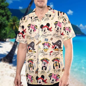May include: Short-sleeved button-down shirt with a beige background featuring cartoon pirate characters including Mickey Mouse, Minnie Mouse, Donald Duck, Daisy Duck, Pluto, and Goofy. The shirt has a pirate theme with pirate hats, swords, and treasure map elements.