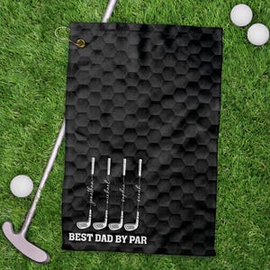 May include: Black golf towel with a honeycomb pattern, golf club illustrations, and the text "BEST DAD BY PAR." Names are printed below each golf club. Golf balls and a putter are also visible on the green grass.