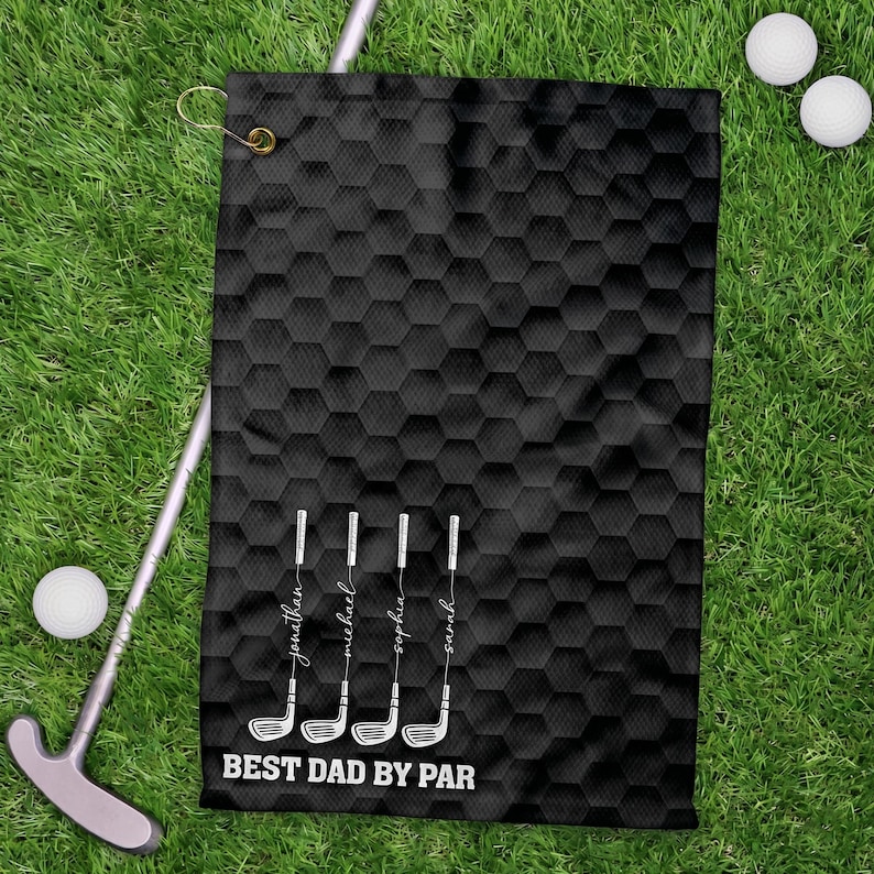 May include: Black golf towel with a honeycomb pattern, golf club illustrations, and the text "BEST DAD BY PAR." Names are printed below each golf club. Golf balls and a putter are also visible on the green grass.