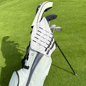May include: A white golf towel featuring a golf club design and the words "BEST GRANDPA". The towel is draped over a golf bag filled with clubs. The names of family members are printed on the towel.
