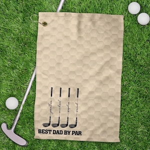 May include: Beige golf towel with a honeycomb design, displaying four golf club illustrations and names: Jonathan, Michael, Sophia, and Sarah. The text "BEST DAD BY PAR" is printed below. Golf balls and a putter are on the green grass.