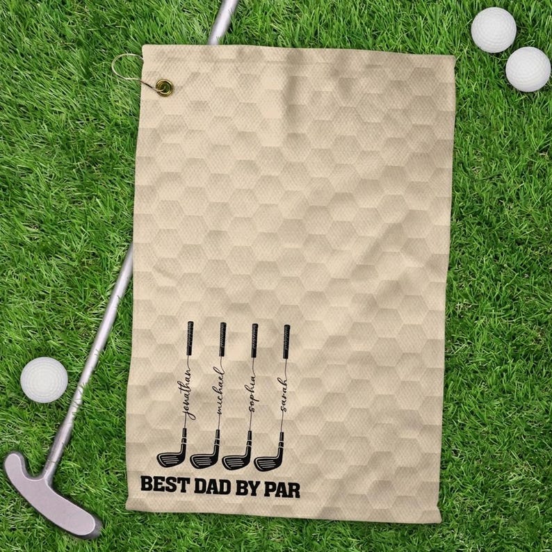 May include: Beige golf towel with a honeycomb design, displaying four golf club illustrations and names: Jonathan, Michael, Sophia, and Sarah. The text "BEST DAD BY PAR" is printed below. Golf balls and a putter are on the green grass.