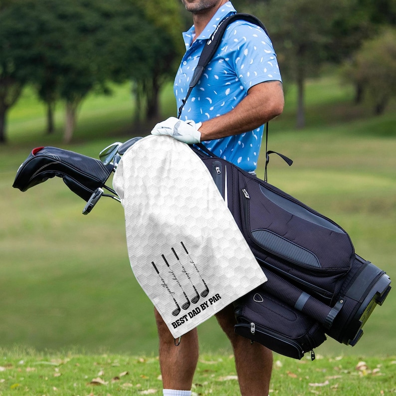 May include: A white golf towel with a honeycomb pattern and black golf club illustrations. The towel has the text "BEST DAD BY PAR" printed on it. The towel is draped over a golf bag.
