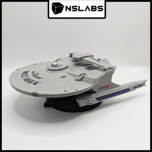 Puede incluir: Modelo gris de la nave estelar USS Reliant de Star Trek, con el texto "NCC-1864" visible en la sección del platillo. El modelo está sobre una base negra y presenta detalles en azul y rojo.