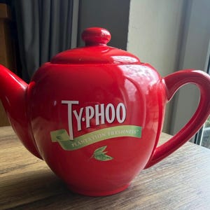 May include: A bright red ceramic teapot with a rounded body, a curved handle, and a matching lid. The front features the white text "TYPHOO" above a green ribbon graphic with the text "PLANTATION FRESHNESS" and a small green tea leaf illustration.