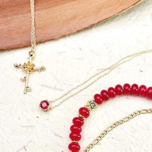 May include: Gold-toned necklaces and a bracelet are displayed on a textured surface. One necklace features a cross pendant with a floral design. Another has a red gemstone pendant. The bracelet has red beads and gold accents.