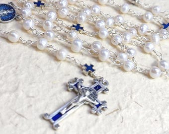 Può includere: Un rosario con perle d'acqua dolce bianche collegate da filo d'argento, intervallate da piccoli accenti a croce in smalto blu. Il rosario include un crocifisso in argento con dettagli in smalto blu e una medaglia circolare in smalto blu.