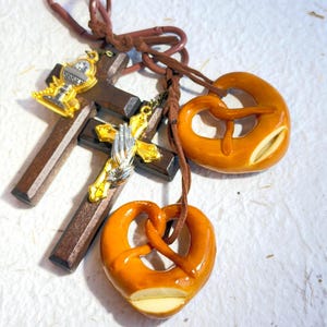 May include: A collection of religious and food-themed charms. Includes two brown wooden crosses with gold and silver religious symbols, and two pretzel-shaped charms in shades of brown and tan, all attached to brown cords.