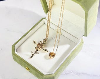May include: Two gold necklaces displayed in an open, green velvet jewelry box. One necklace features a floral pendant, the other a hexagonal gemstone. The box is lined with white fabric, set against a marble surface.