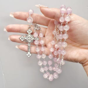 Può includere: Un rosario con perline lisce e traslucide rosa chiaro collegate da catene in metallo argentato. Il rosario include un crocifisso argentato con dettagli in smalto bianco e una figura centrale, collegato alle perline da un anello circolare.