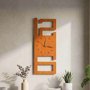 Op de afbeelding: Een houten wandklok met een uniek rechthoekig ontwerp. De klok is gemaakt van lichtbruin hout en heeft een minimalistische stijl met een zichtbare wijzerplaat en wijzers. De klok hangt aan een lichtgrijze muur.
