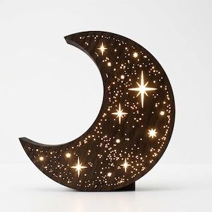 Puede incluir: Una lámpara de noche de madera con forma de media luna y diseño de estrellas. La luna marrón oscuro presenta estrellas recortadas de varios tamaños que emiten un brillo cálido. La lámpara es independiente y se apoya en una pequeña base.