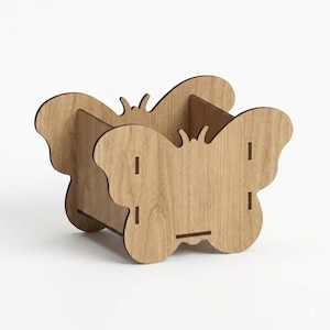 Puede incluir: Un organizador de madera con forma de mariposa y acabado marrón claro. El organizador tiene un compartimento rectangular y está construido con varias piezas de madera. Las alas de mariposa son visibles en los laterales, y el diseño es sencillo y elegante.