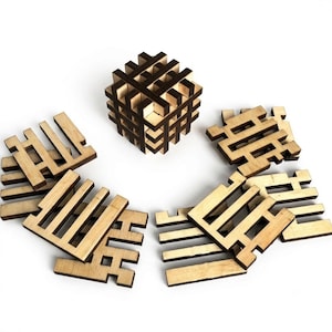 Puede incluir: Un cubo rompecabezas de madera ensamblado a partir de piezas entrelazadas, acompañado por varios componentes de madera planos y de color claro con bordes dentados dispuestos en un patrón semicircular alrededor del cubo central.