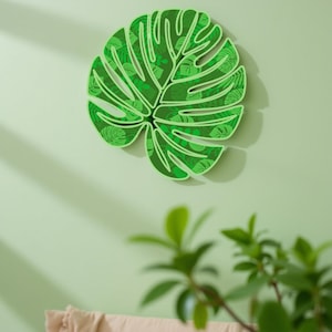 Puede incluir: Decoración de pared verde con forma de hoja, con un patrón detallado de hojas más pequeñas. La decoración está montada en una pared verde claro, creando un tema tropical o botánico. El diseño es de un tono verde vibrante.