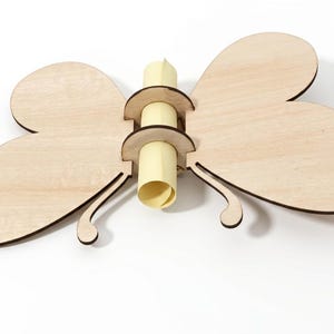 Op de afbeelding: Een houten vlindervormige houder met een rol. De vlinder is gemaakt van lichtgekleurd hout met donkerbruine randen. De rol is lichtgeel van kleur en wordt op zijn plaats gehouden door twee cirkelvormige houten stukken.