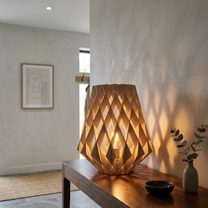 Op de afbeelding: Een houten tafellamp met een geometrisch ruitpatroon. De lamp is gemaakt van lichtgekleurd hout en staat op een donkere houten tafel. Een kleine zwarte kom en een vaas met groen staan ook op de tafel.