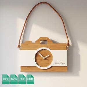 Puede incluir: Un reloj de pared de madera con forma de cámara con una franja blanca y una esfera redonda. El reloj tiene una correa de cuero marrón para colgarlo. El texto "Amee House" está en la esfera.