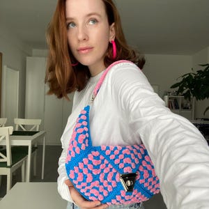 Könnte beinhalten: Eine blau-rosa gehäkelte Handtasche mit silbernem Verschluss. Die Tasche hat ein geometrisches Muster aus blauen und rosa Quadraten. Der Riemen ist rosa. Die Person trägt ein weißes Hemd und blaue Jeans.