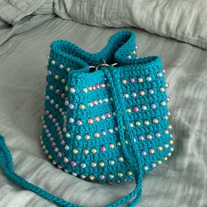 May include: A turquoise crochet bucket bag adorned with iridescent beads. The handmade bag features a drawstring closure and a matching turquoise strap. The beads are in various pastel colors, adding a touch of sparkle to the bag's textured surface.