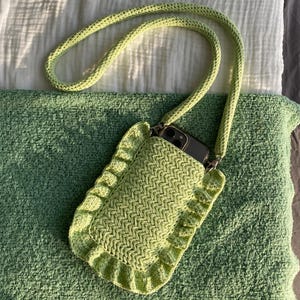 May include: A light green crochet phone bag with a matching strap. The bag has a rectangular shape with a ruffled edge. The phone is visible inside the bag. The bag is lying on a green textured surface.