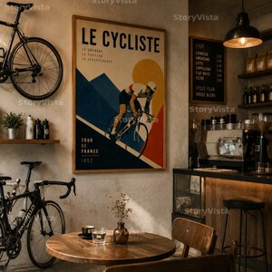 May include: A vintage-style poster titled "Le Cycliste" featuring a cyclist in action, with text in French. The poster is framed and hangs in a cafe setting, alongside a bicycle and coffee menu. The cafe has a wooden table and chairs.
