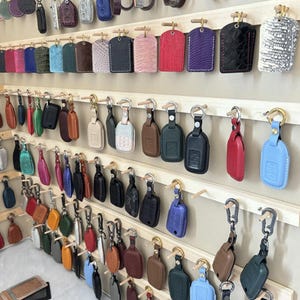 Puede incluir: Exhibición de varios llaveros de cuero en múltiples colores y texturas. Los llaveros están colgados de varillas de madera, mostrando una variedad de diseños, incluyendo colores sólidos, patrones en relieve y diferentes formas. Los llaveros están sujetos a anillos metálicos.