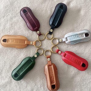 Puede incluir: Colección de siete fundas de cuero para llaveros en varios colores, incluyendo burdeos, negro, beige, plateado, rojo, verde y marrón. Cada funda está unida a un llavero dorado.