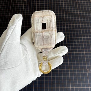 May include: A cream leather key fob cover with a textured pattern, featuring a rectangular opening and a small strap with a gold-coloured ring. The fob is held in a white glove.