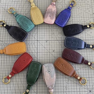 Puede incluir: Una colección de llaveros de cuero en varios colores, incluyendo negro, azul, verde y rojo. Cada llavero presenta un diseño de llave Mercedes-Benz con un anillo dorado para sujetar.