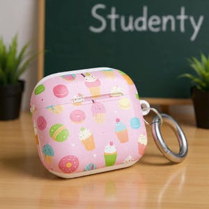 Puede incluir: Funda rosa para auriculares inalámbricos con un estampado de coloridos cupcakes, donuts y macarons. La funda tiene un mosquetón plateado. La palabra "Studenty" está escrita en blanco sobre una pizarra verde en el fondo.