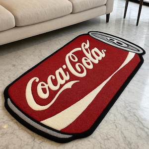 Pode incluir: Um tapete vermelho em forma de lata de Coca-Cola com letras brancas e uma faixa branca. O tapete tem uma borda preta e um topo prateado, assemelhando-se a uma lata de refrigerante. O tapete está colocado num chão de mármore.