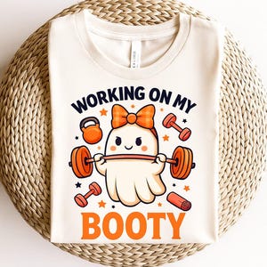 May include: Cream-colored t-shirt with a cartoon ghost lifting weights. The shirt features the text "WORKING ON MY BOOTY" in navy and orange. Additional orange dumbbells, a kettlebell, and stars are also on the shirt.