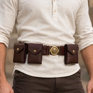 Peut inclure: Une ceinture utilitaire en cuir marron foncé dotée de trois pochettes rectangulaires avec fermetures à pression en laiton et d'une boucle centrale circulaire en laiton ornée d'un emblème ailé, portée sur une chemise crème à manches longues.