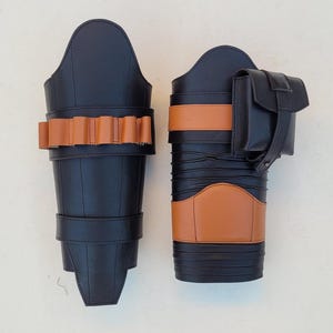 May include: Two black leather leg armor pieces with tan accents. One piece features a series of tan-colored loops, while the other has a tan band and a small black pouch. The armor is designed with a sleek, modern aesthetic.