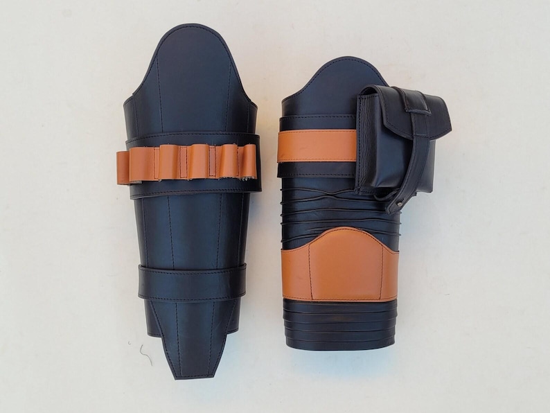 May include: Two black leather leg armor pieces with tan accents. One piece features a series of tan-colored loops, while the other has a tan band and a small black pouch. The armor is designed with a sleek, modern aesthetic.