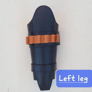 May include: A black leather leg armor with a brown leather band of bullet holders and a strap. The text "Left leg" is in a blue box at the bottom. The armor is designed to protect the leg.