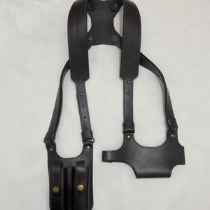 以下が含まれることがあります： 2つのマガジンポーチと別のホルスターコンポーネントを備えた、濃い茶色の革製ショルダーホルスター。 ホルスターは調節可能なストラップと真鍮色の金具を備えています。 デザインは胸と背中に着用することを目的としています。