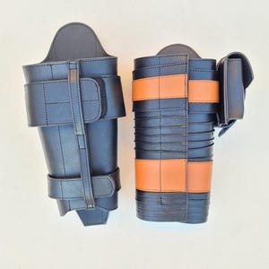 May include: Two black leather leg armor pieces. One has a simple design with straps and buckles. The other features orange bands and a small pouch. Both are likely for costume or historical reenactment.