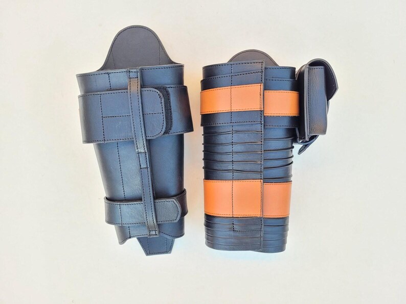 May include: Two black leather leg armor pieces. One has a simple design with straps and buckles. The other features orange bands and a small pouch. Both are likely for costume or historical reenactment.