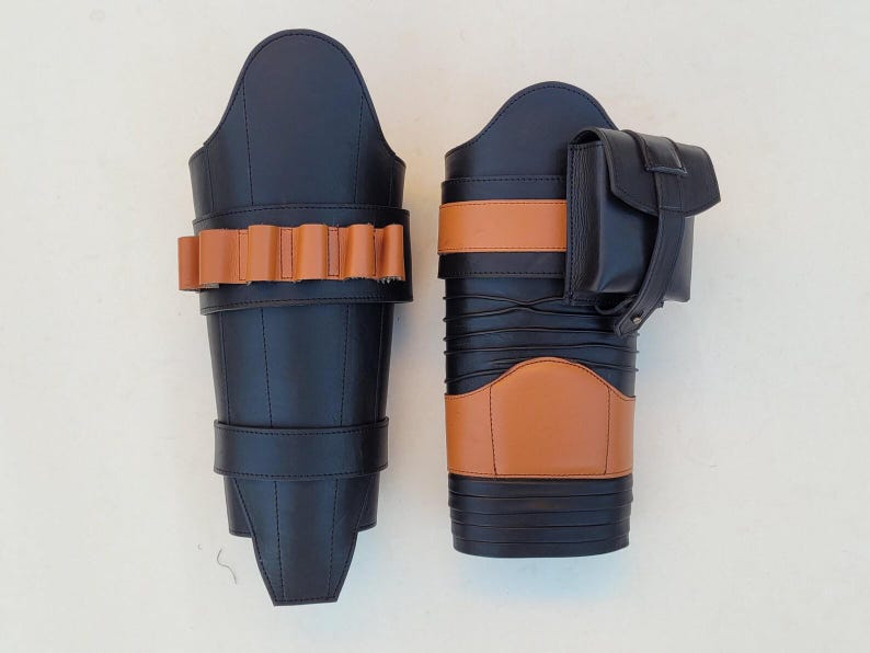 May include: Two black leather leg armor pieces with brown accents. One piece features a row of brown cartridge holders, while the other has a small pouch. The armor is designed with multiple straps and panels.