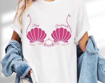 May include: White t-shirt with a pink graphic design featuring seashell shapes, the words "Bride" and "Bachelorette", the city "Miami", and the date "July 2025". The shirt has a round neckline and short sleeves.