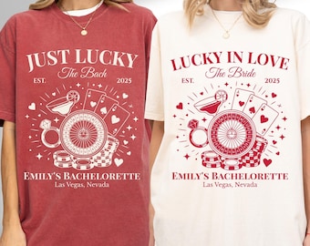 May include: Two t-shirts, one crimson and one ivory, feature a casino-themed design with the text "JUST LUCKY The Bach 2025" and "LUCKY IN LOVE The Bride 2025". The design includes a roulette wheel, playing cards, and the text "EMILY'S BACHELORETTE Las Vegas, Nevada".